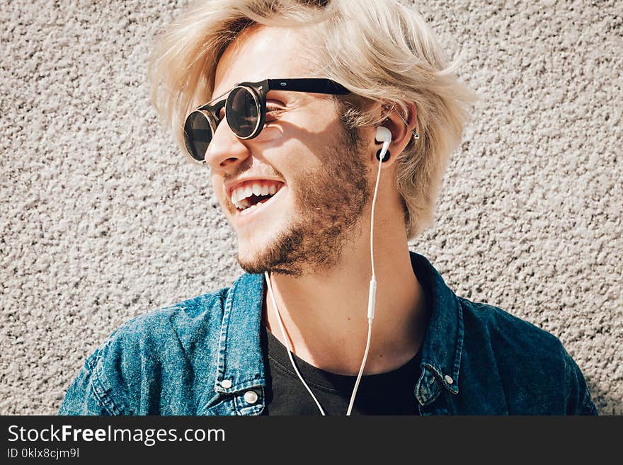 Men fashion, technology, urban style clothing concept. Hipster smiling guy standing on city street wearing jeans outfit listening to music through earphones. Men fashion, technology, urban style clothing concept. Hipster smiling guy standing on city street wearing jeans outfit listening to music through earphones