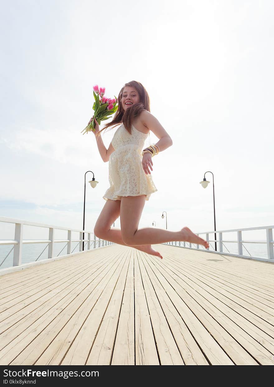 Happiness, romance, success concept. Woman wearing short white dress jumping on pier holding bouquet of flowers. Happiness, romance, success concept. Woman wearing short white dress jumping on pier holding bouquet of flowers.