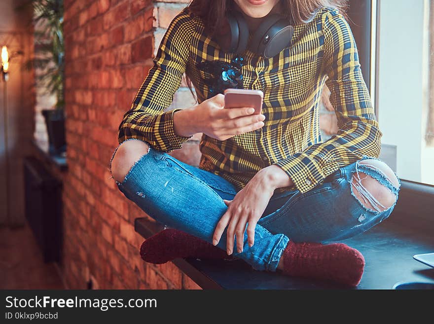 Cropped image of a student girl in casual clothes listening music while using a smartphone while sitting on a window