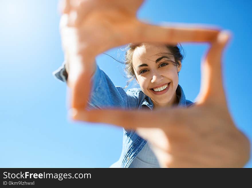 Portrait of a young smiling attractive woman in jeans clothes at sunny day on the blue sky background. woman shows a frame from hands like photo. Photo Frame Hands Made By A Hipster young girl. Portrait of a young smiling attractive woman in jeans clothes at sunny day on the blue sky background. woman shows a frame from hands like photo. Photo Frame Hands Made By A Hipster young girl