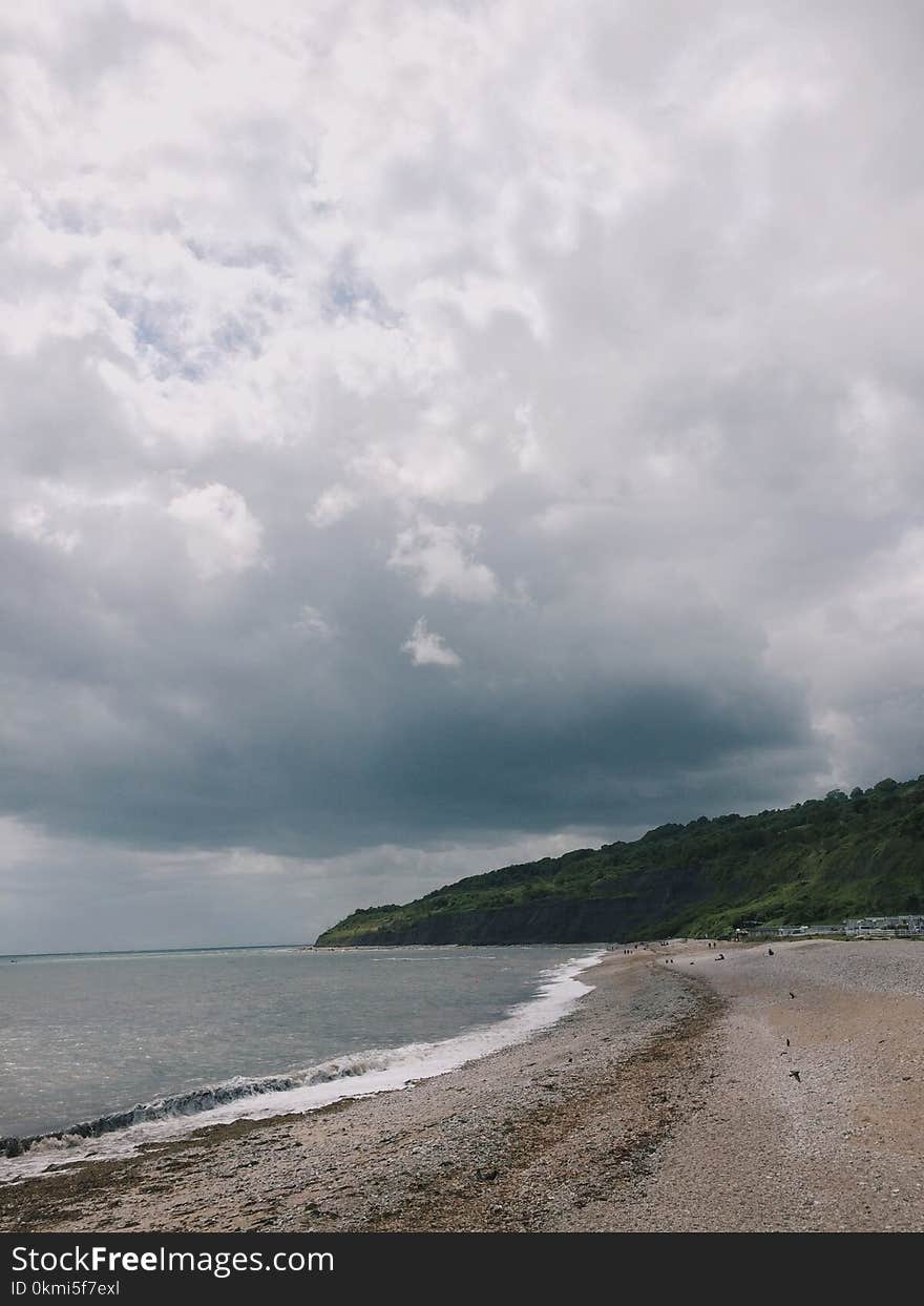 Beach Sand Near Body of Water and Green Mountain Under Cloudy Sky