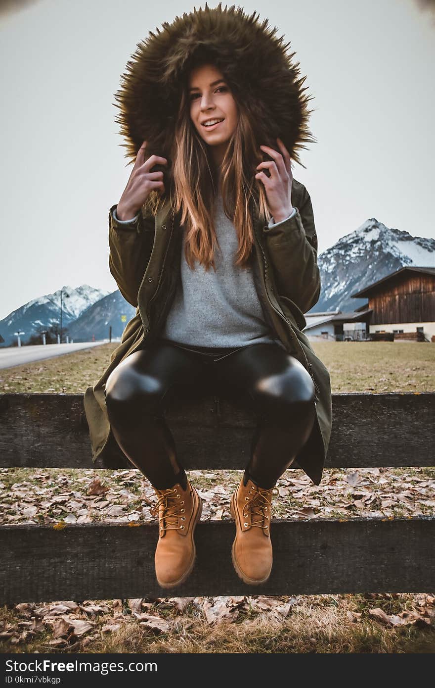 Woman Wearing Brown and Green Parka Sitting on Gray Wooden Fence at Daytime