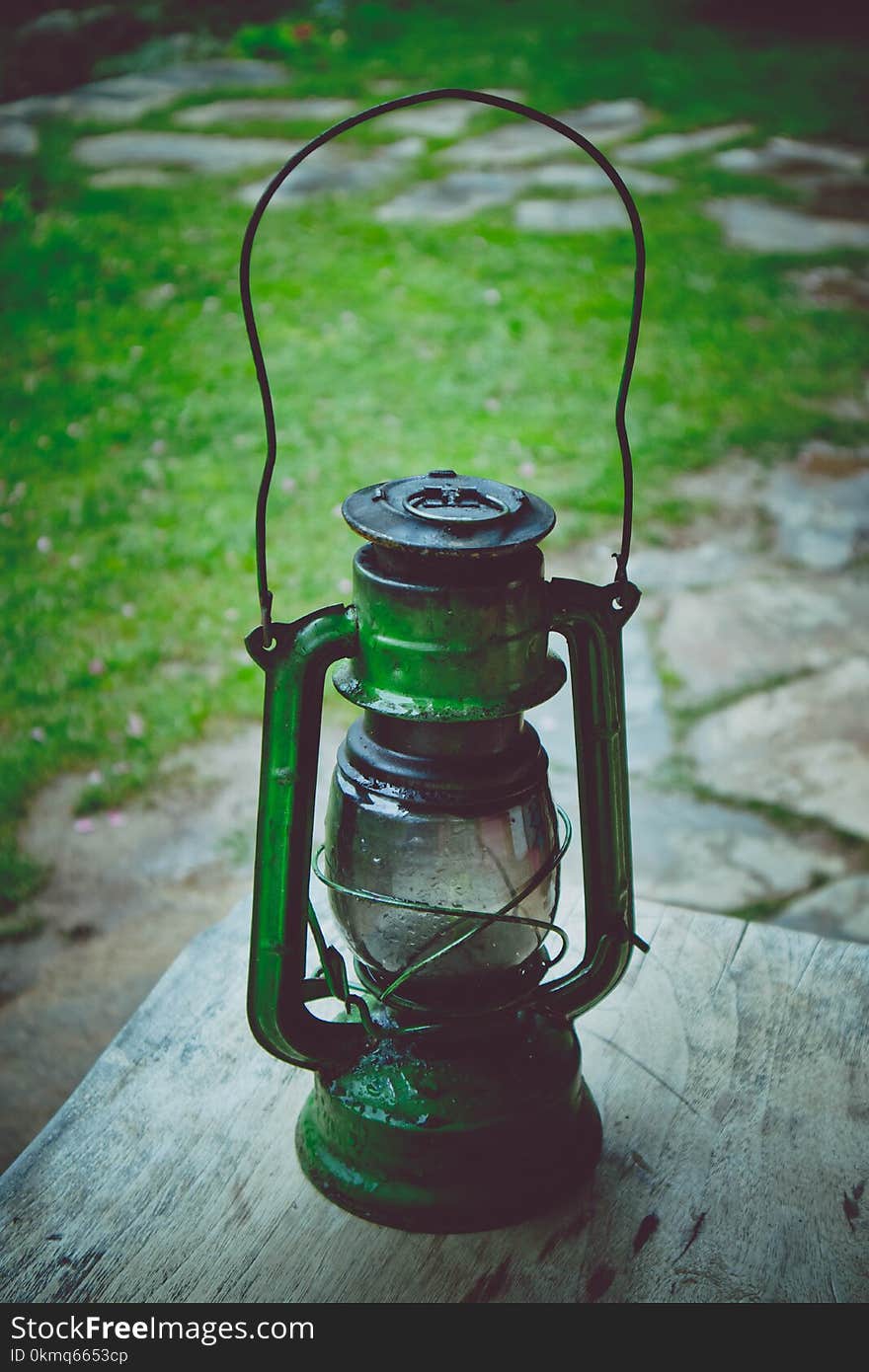 Photo depicts metal rusty lantern. An old oil kerosene green lampplaced on a wooden table in the garden. Summertime, footpath paved with stone and blurred nature on the background.