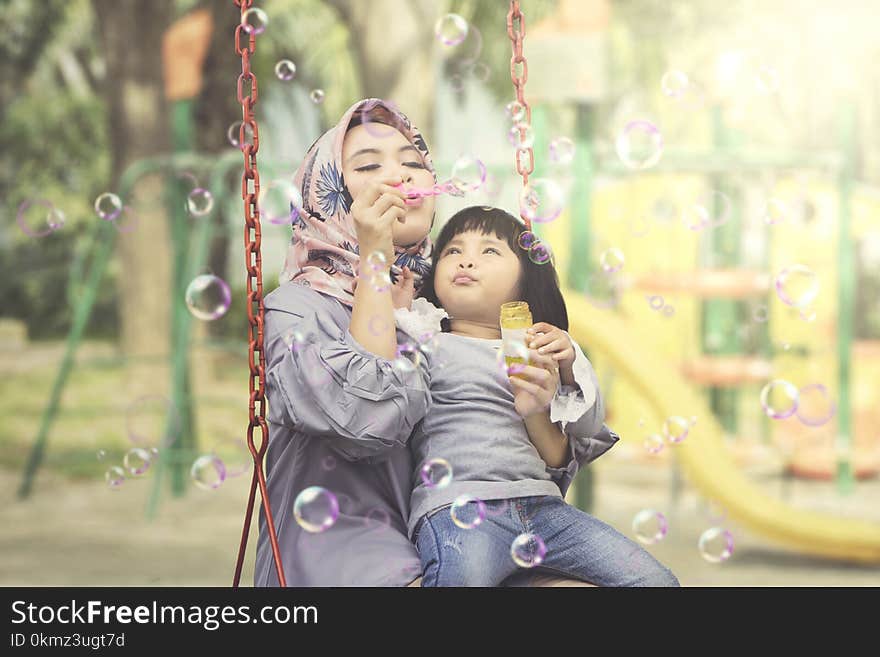 Image of a Muslim women with her daughter blowing soap bubbles while sitting on the swing. Shot in the playground. Image of a Muslim women with her daughter blowing soap bubbles while sitting on the swing. Shot in the playground