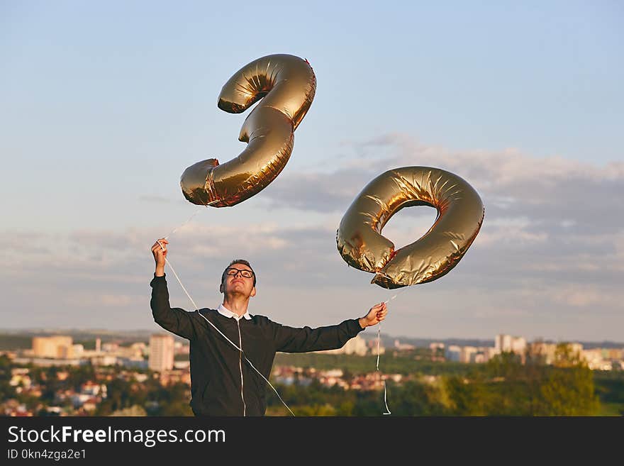 Man celebrates thirty years birthday. Person holding helium balloons in shape of number 30 against city.