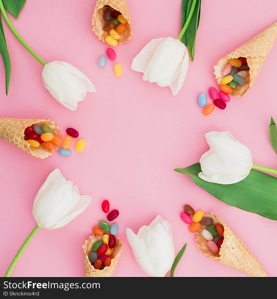 Frame made of sugar candy in waffle cones and white tulip flowers on pink background. Flat lay, top view