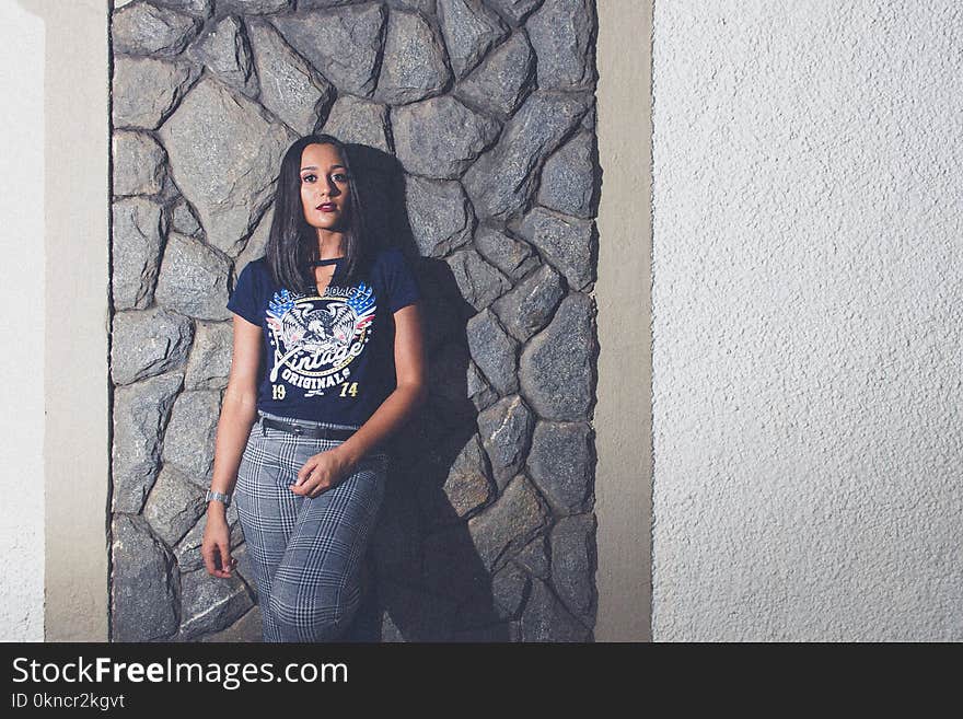 Woman Wearing Blue Wings-printed V-neck T-shirt and Gray Pants Leaning on Gray Rock Cladding