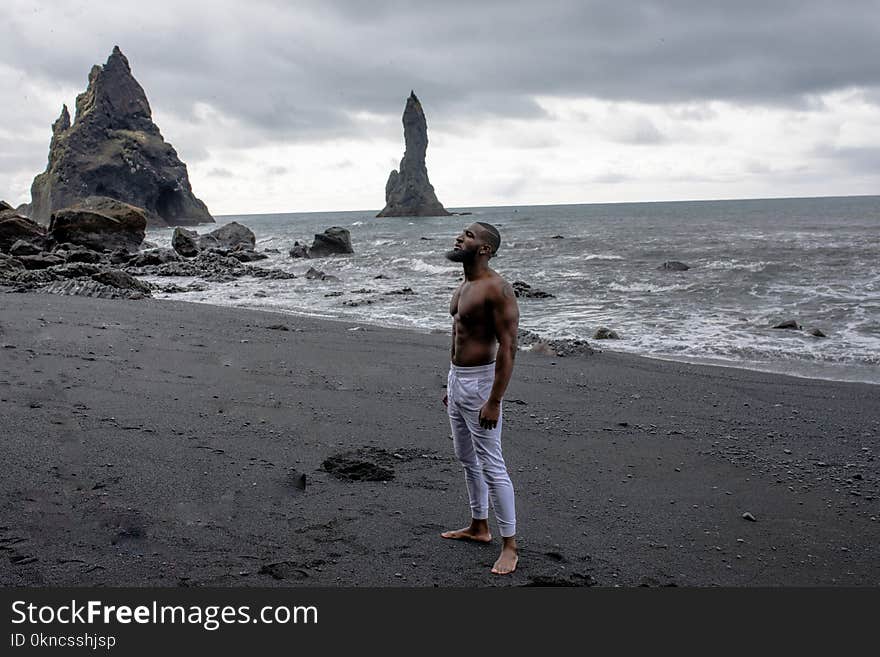 Man in White Jeans Stands on Grey Sand in Beach Under Grey Clouds