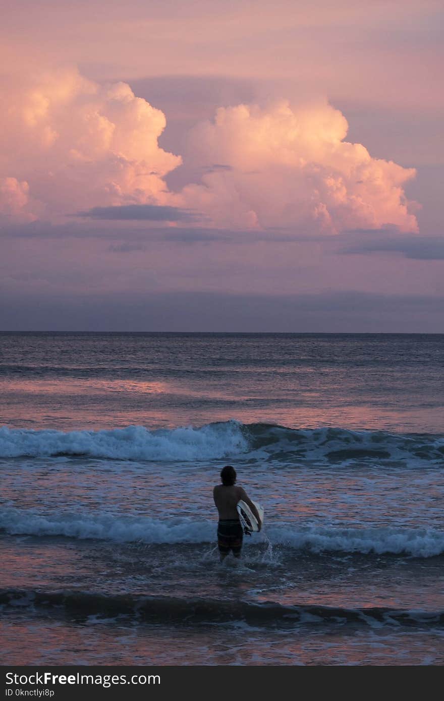 Person Carrying Surfboard Walking Against Sea Waves during Golden Hour