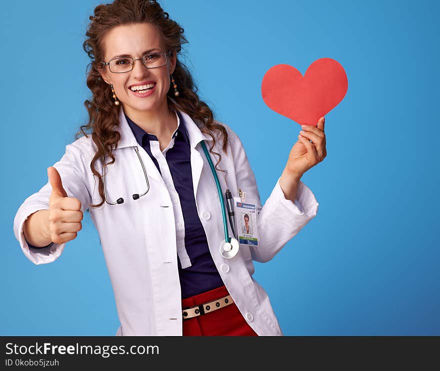 Smiling medical practitioner woman in white medical robe showing thumbs up and heart isolated on blue background. Smiling medical practitioner woman in white medical robe showing thumbs up and heart isolated on blue background