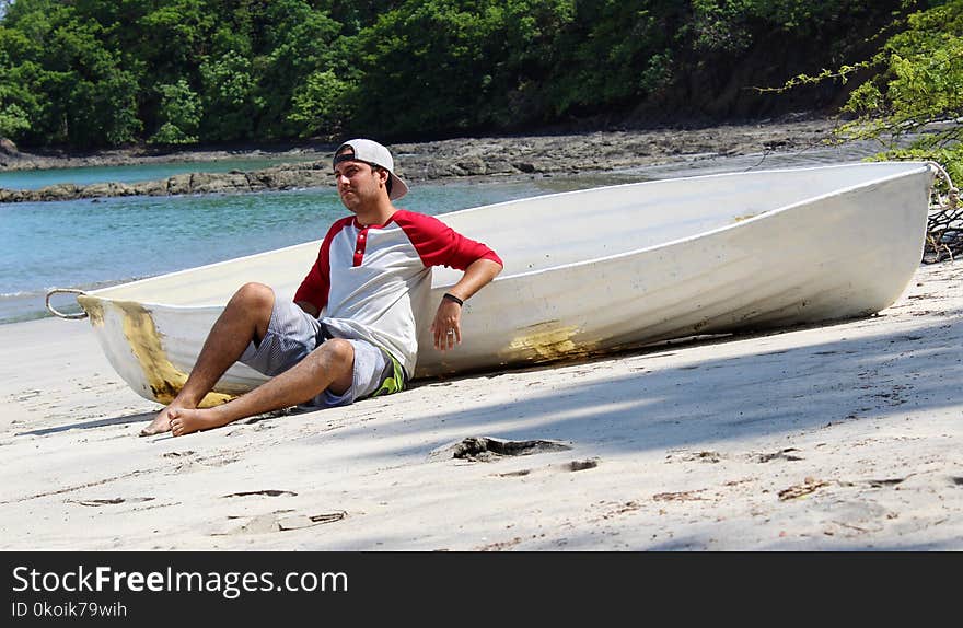 Good looking castaway man sitting in the beach by a wrecked boat waiting for help with ocean and jungle in the background.