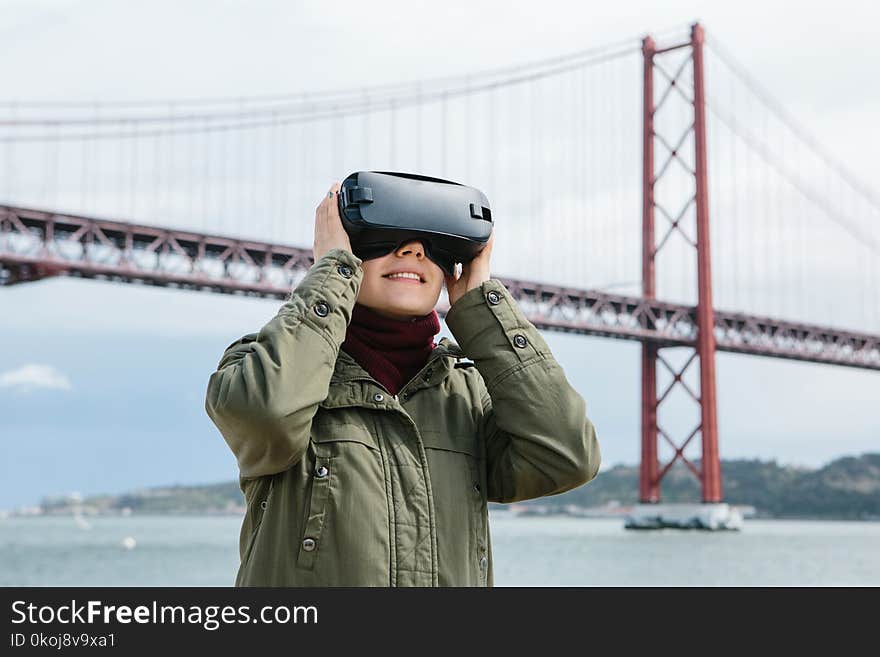 Young beautiful girl wearing virtual reality glasses. 25th of April bridge in Lisbon in the background. The concept of modern technologies and their use in everyday life.