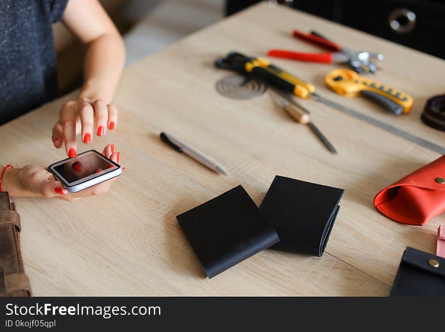 Female hands with red nails at leather atelier using smartphone, handmade wallets and tools on table. Concept of handicraft business and craftswoman with modern technology.