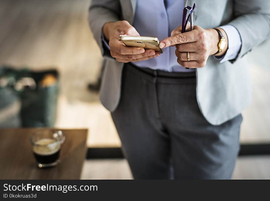 Person Wearing Grey Suit Jacket And Black Bottoms Standing Near Brown Wooden Table Holding Smartphone