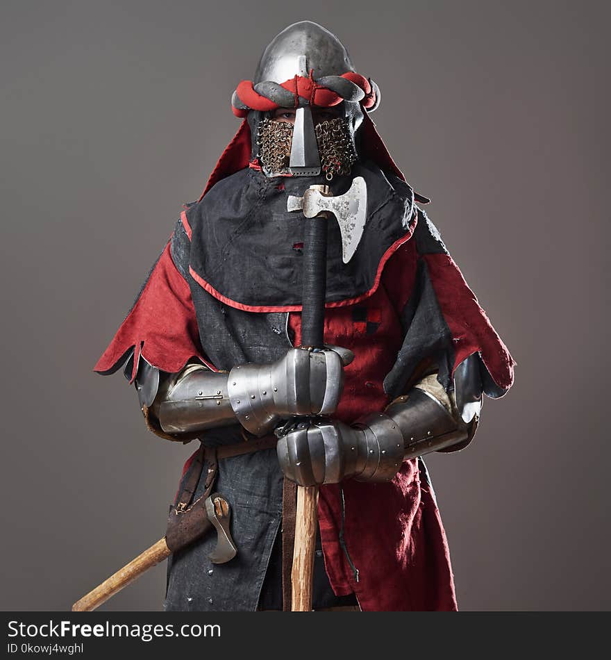 Medieval knight on grey background. Portrait of brutal dirty face warrior with chain mail armour red and black clothes and battle axe.