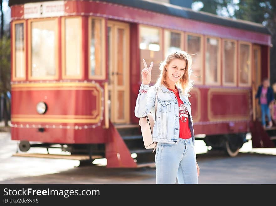 Beautiful girl in a blue denim jacket and jeans against a red tram. Woman shows the world with gestures in the sunset sun with a backpack on the bus background