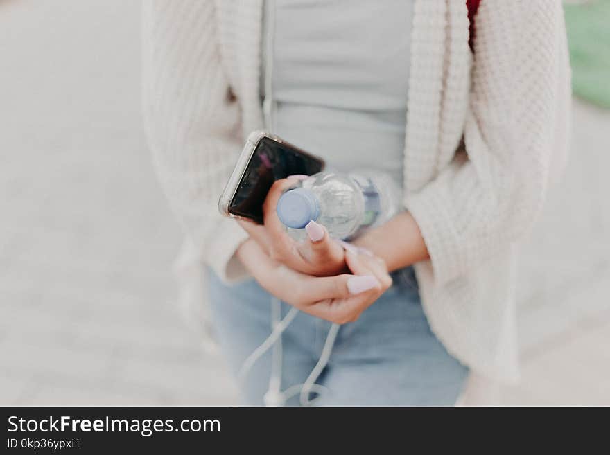 Selective Focus Photography of Woman in White Cardigan Holding Water Bottle and Black Smartphone