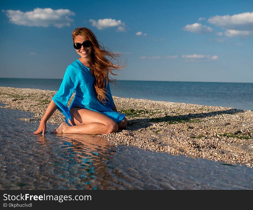 Young happy woman in sunglasses, blue tunika sits at the seaside, laugh and looks at the sea at sunny day. Beautiful Smiling Woman Laughing On Summer Travel Vacation. Relationships, Summertime. Young happy woman in sunglasses, blue tunika sits at the seaside, laugh and looks at the sea at sunny day. Beautiful Smiling Woman Laughing On Summer Travel Vacation. Relationships, Summertime