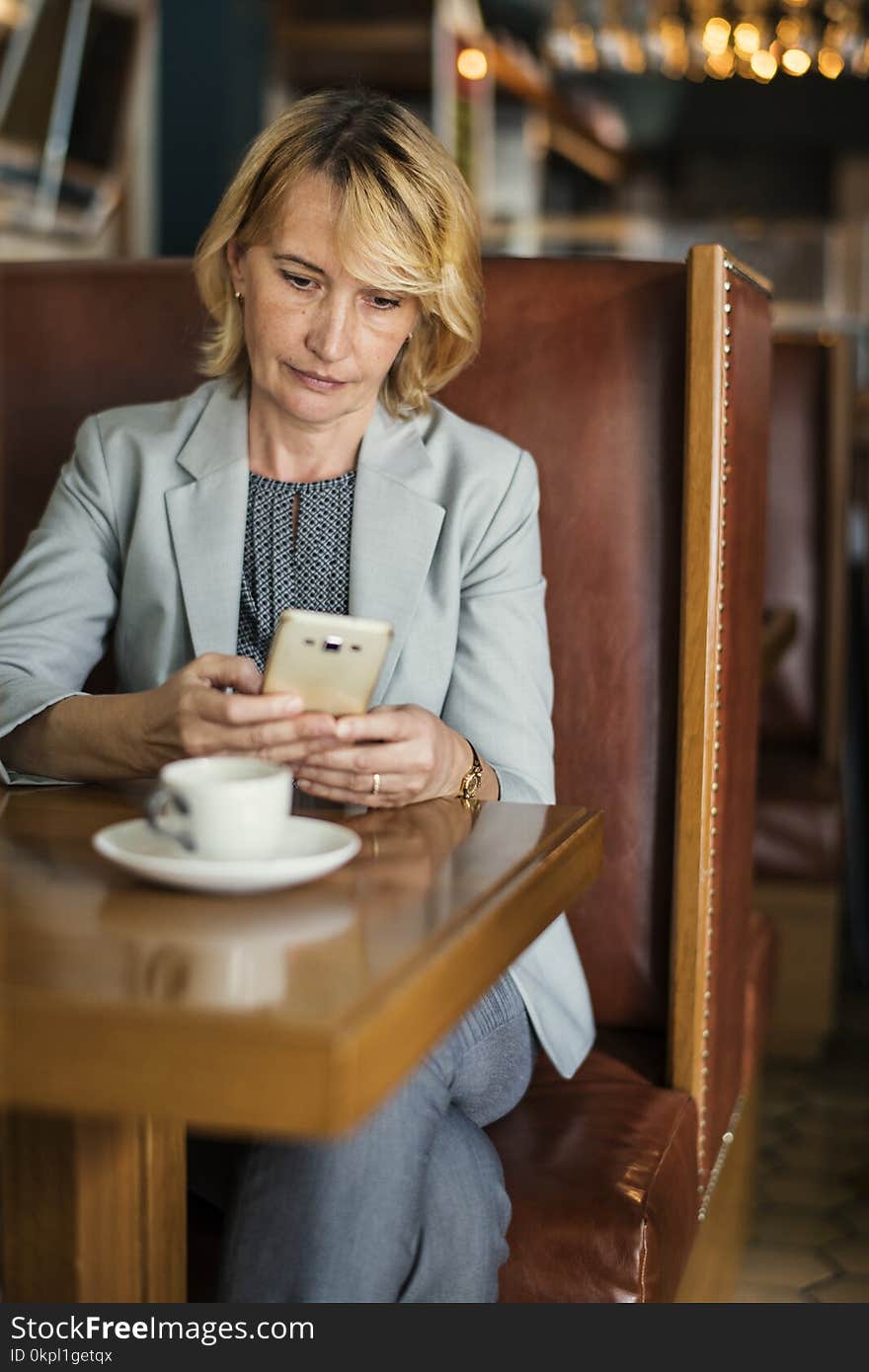 Woman in Grey Notched Lapel Suit Jacket Holding Smartphone Sitting Beside Wooden Table