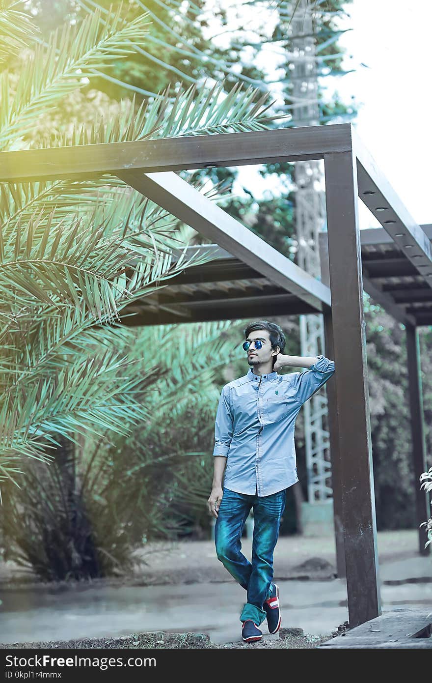 Man Wearing Blue Button-up Long-sleeved Shirt and Sunglasses Leaning on Wooden Canopy