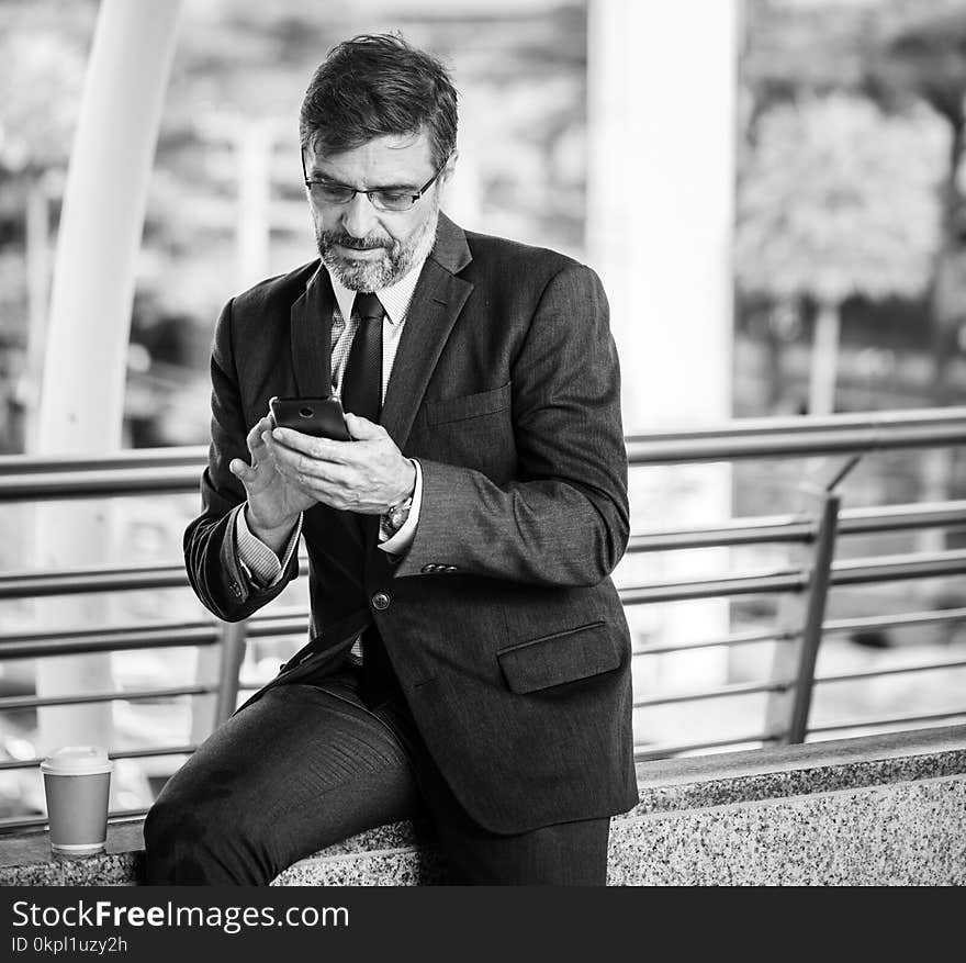 Greyscale Photo Of Man Wearing Suit Jacket And Eyeglasses Holding Smartphone