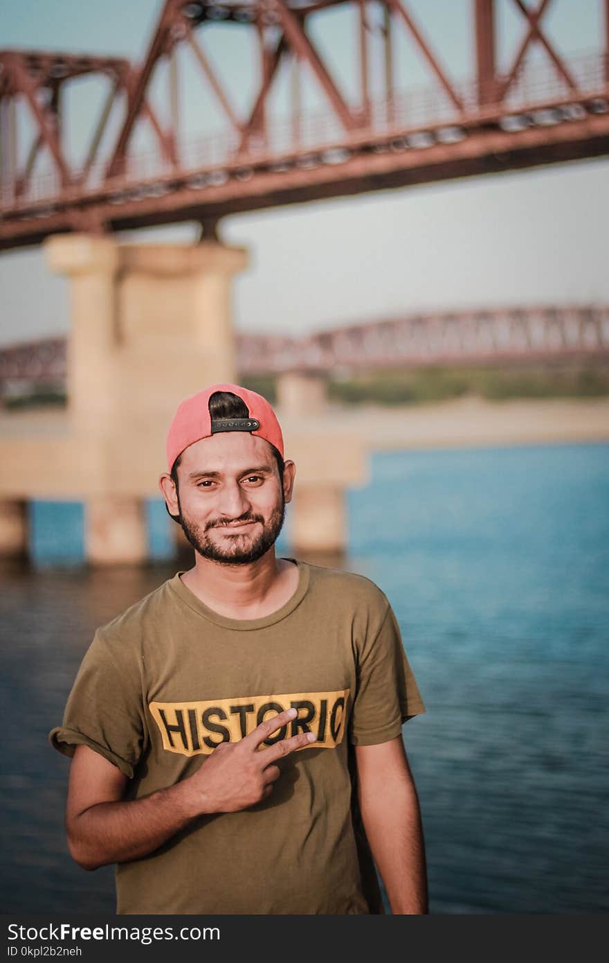 Man Wearing Gray Historic-printed T-shirt and Red Snapback Cap Taking Photo Beside Body of Water