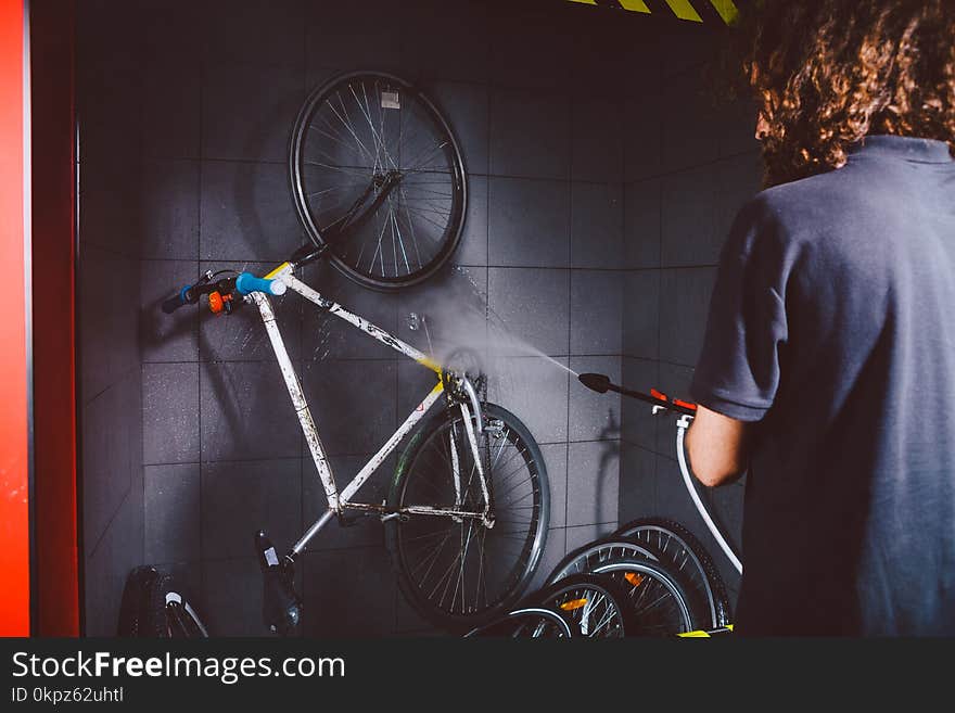 Services professional washing of a bicycle in the workshop. A young caucasian stylish man with long curly hair does a bicycle cleaning using an automatic electric water pump.