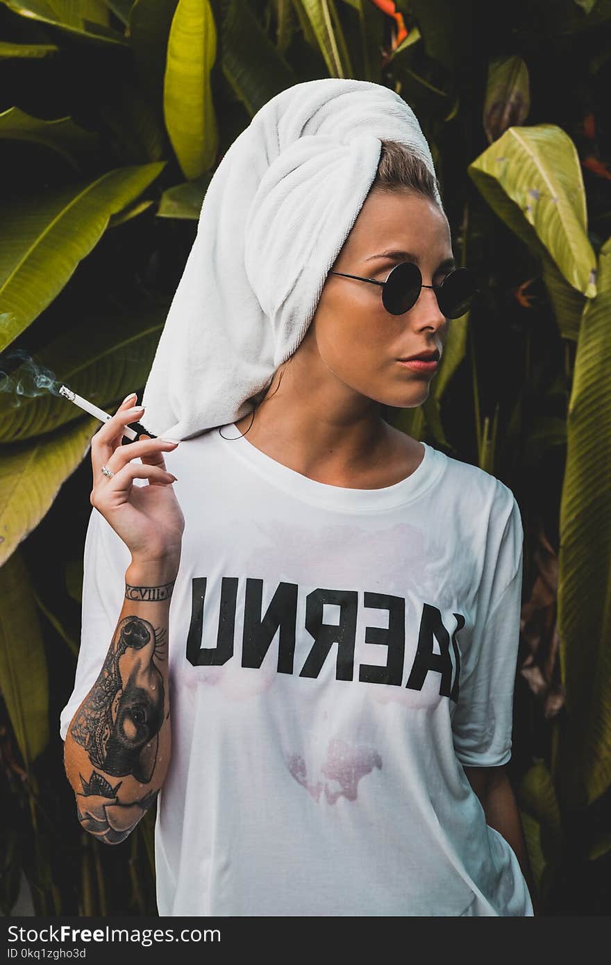Woman Wearing White Crew-neck Shirt and Black Framed Sunglasses With White Bath Towel on Her Head Holding Cigarette Stick