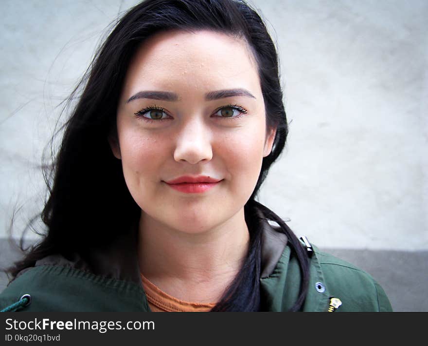 Woman Wearing Green and Black Top Standing Near White Wall Smiling in Front of Camera