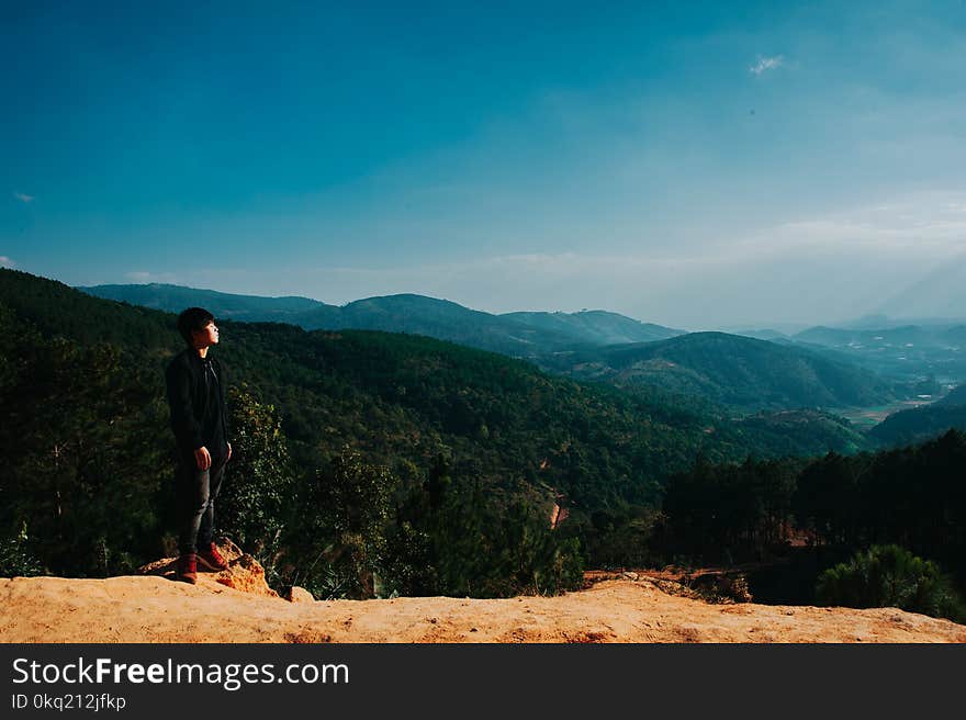 Man Stands on Cliff With Green Mountains on Background