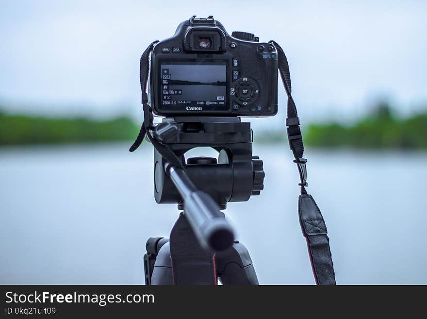 Selective Focus Photo of Black Canon Camera on Tripod Stand in Front of Body of Water Photo