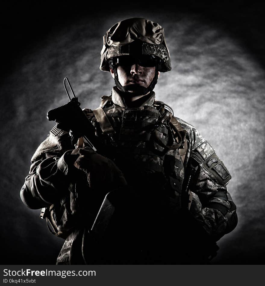 Low key studio portrait of private security service contractor, army infantry rifleman, US marine raider in helmet, sunglasses, camouflage uniform posing with weapon on black background with backlight. Low key studio portrait of private security service contractor, army infantry rifleman, US marine raider in helmet, sunglasses, camouflage uniform posing with weapon on black background with backlight
