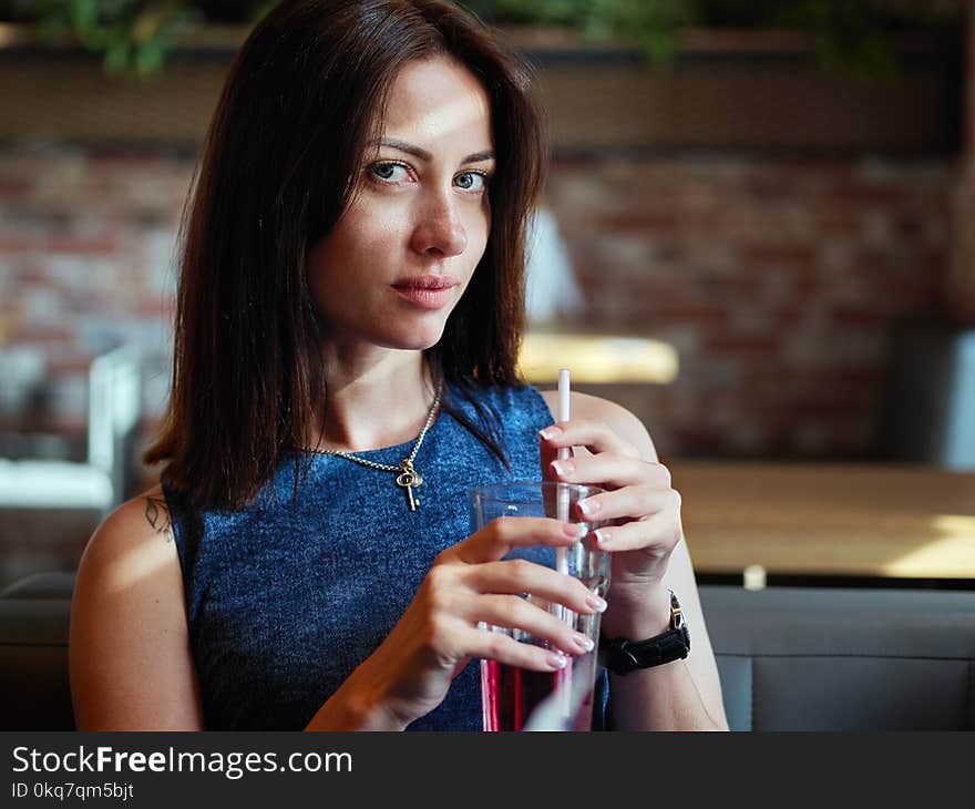 Close up of beautiful adult brunette woman in jeans dress drinking ice tea from glass cup with straw at cafe
