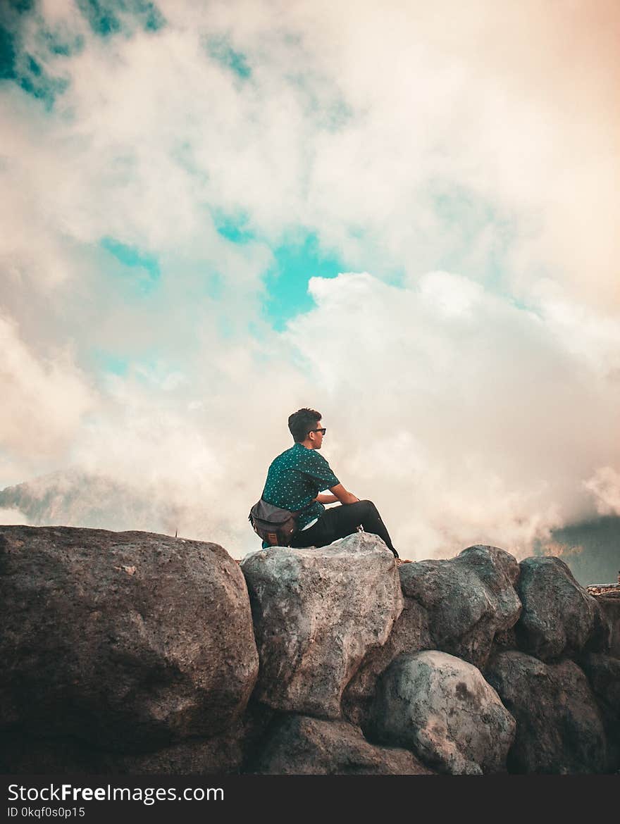 Man in Green Shirt and Black Pants Sitting on Top of Rock Cliff Under White Clouds