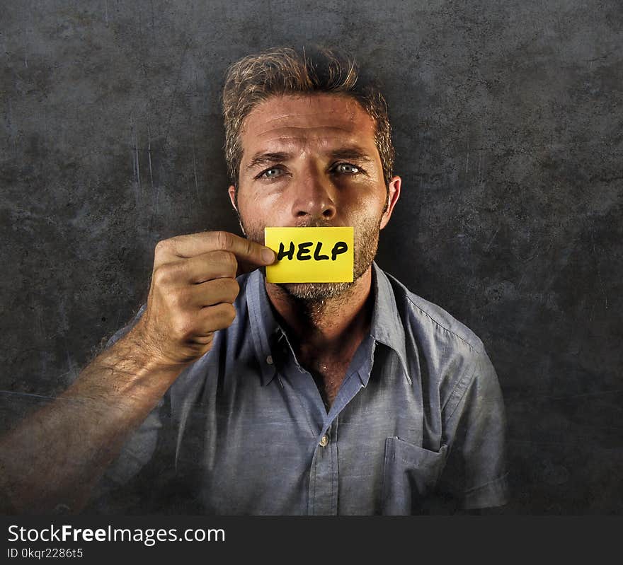 Dramatic portrait of young handsome and depressed man holding paper asking for help feeling sad looking miserable and frustrated suffering depression isolated on black grunge background