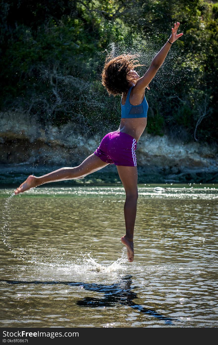 Woman in Blue Sports Bra and Purple Shorts Leaping Above Body of Water