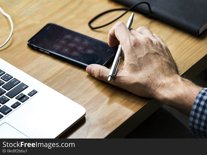 Person Holding Gray Retractable Pen and Black Iphone on Brown Wooden Table