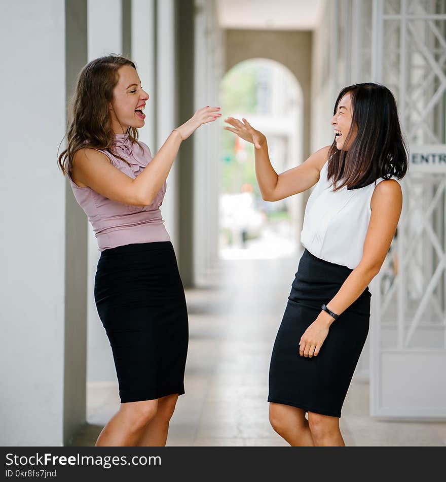 Woman Wearing White Sleeveless Top and Black Pencil Skirt Facing Woman Wearing Pink Sleeveless Top and Black Pencil Skirt Leaning on Wall