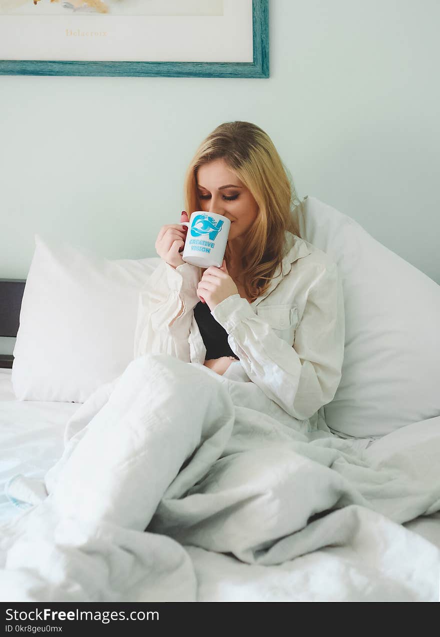 Woman Wearing White Dress Shirt Sitting on Bed While Drinking Through White Ceramic Mug