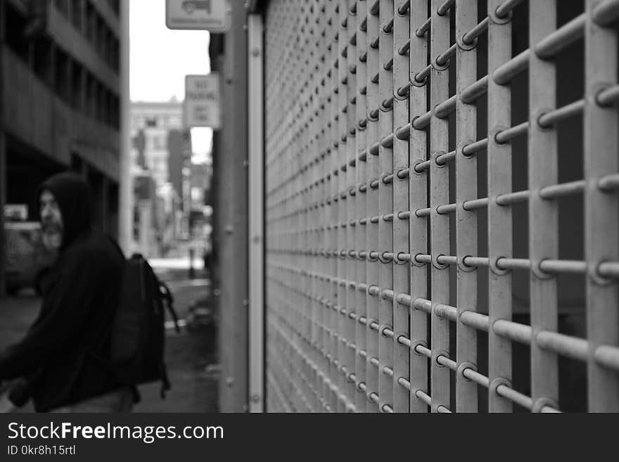 Grayscale Photo of Man in Hoodie in Front of White Window Grill