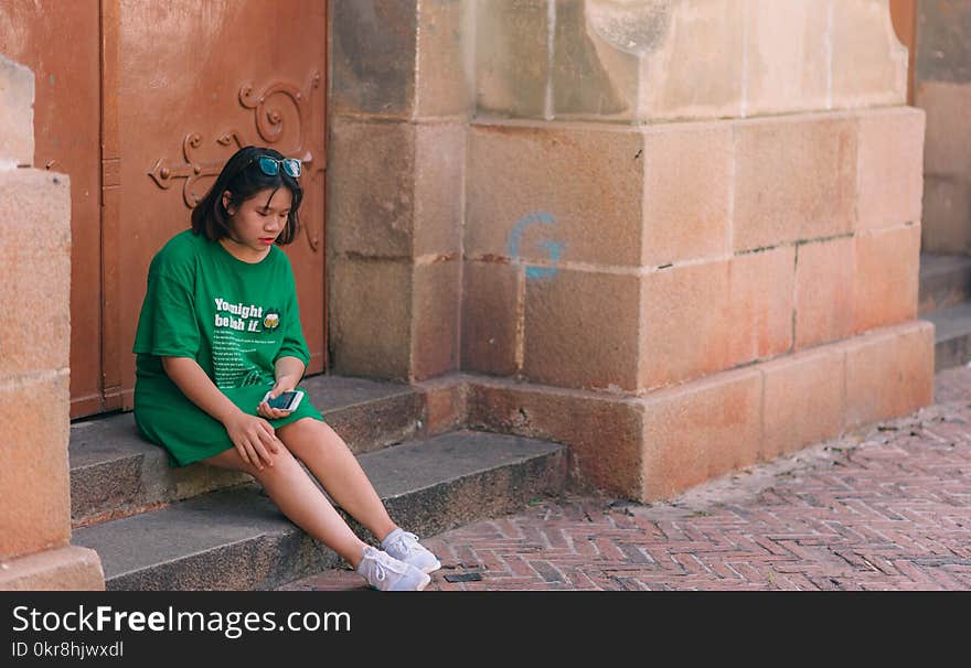 Woman Wearing Green Shirt Sitting Near Brown Gate Holding Smartphone