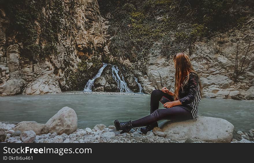 Woman With Brown Hair in Black Jacket Sitting on Rock Near Body of Water