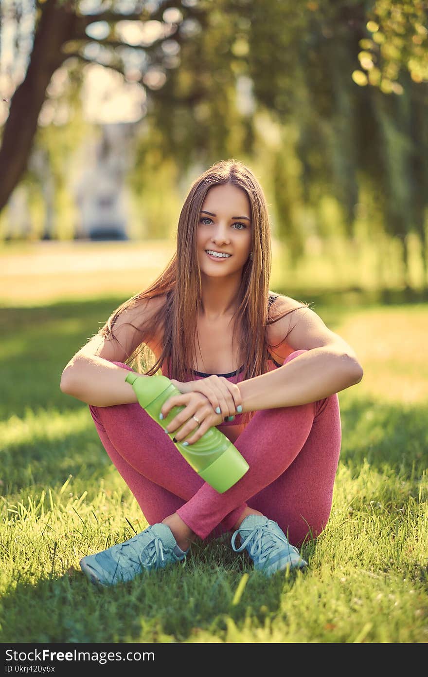 Healthy lifestyle concept. Young girl in sportswear in lotus pose sitting on green grass and holds a bottle of water.n