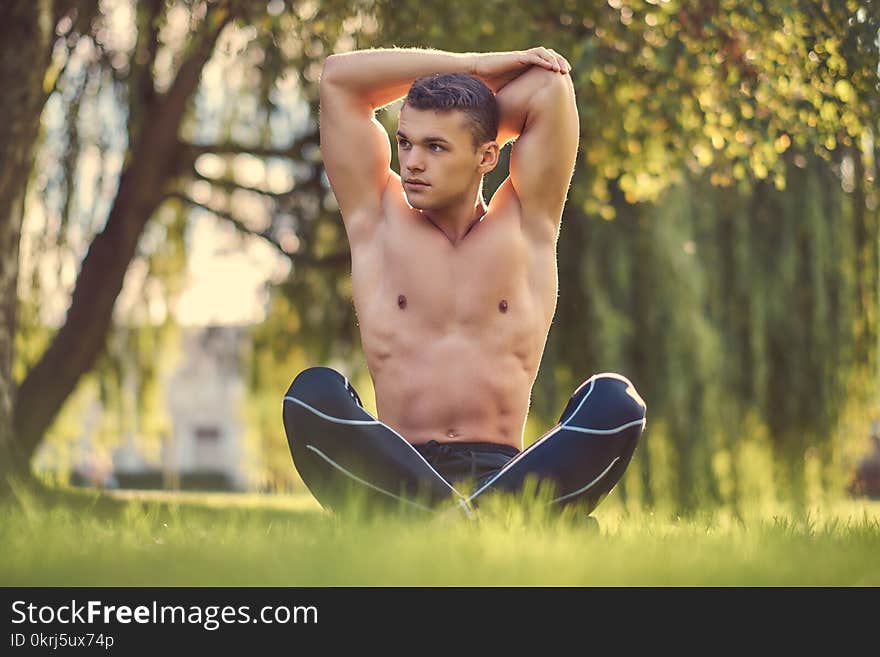 Healthy lifestyle concept. Shirtless young man stretching hands while sitting in lotus pose on a green grass.