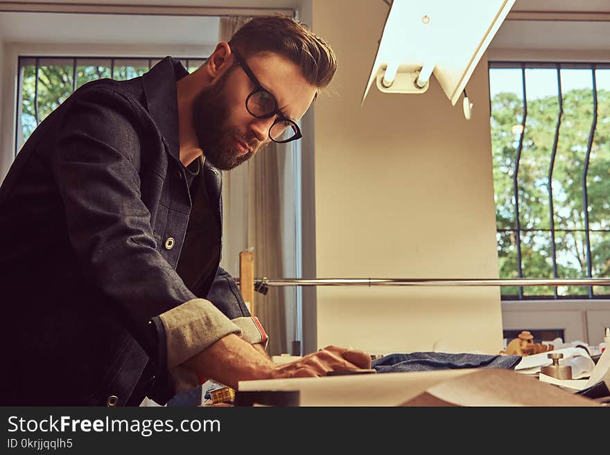 Handsome bearded tailor makes measurements of jeans cloth samples at a sewing workshop.