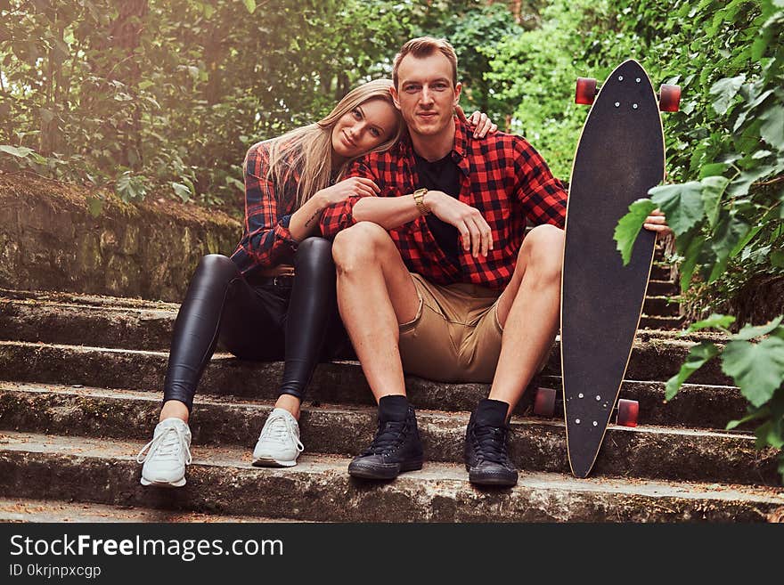 Young hipster couple, handsome men with a skateboard and blonde girl, sitting on steps in the park.