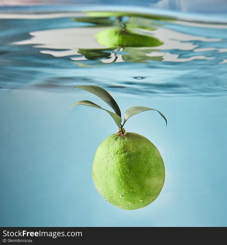 Fresh green lime with leaves falls into the water on a blue background creating waves on the water. The concept of a summer drink. Fresh green lime with leaves falls into the water on a blue background creating waves on the water. The concept of a summer drink