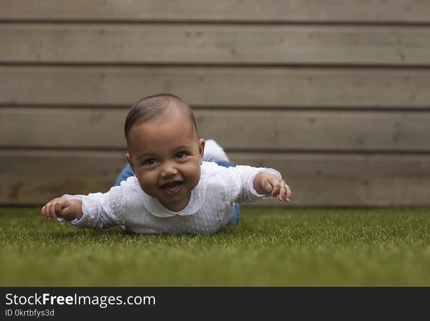 Adorable cute six months old baby girl lying on belly on grass surface with wooden background. Adorable cute six months old baby girl lying on belly on grass surface with wooden background