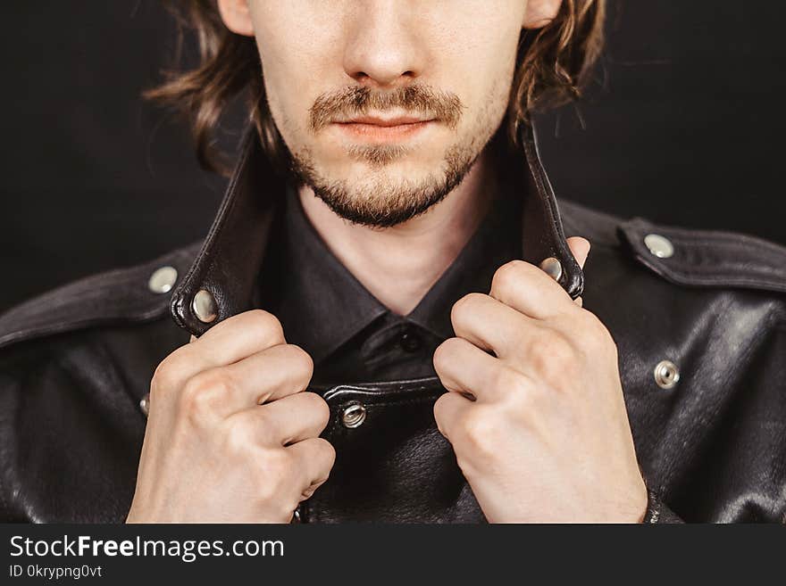 Dramatic fashion model in leather jacket resting elbow on knee while seated on black background. Dramatic fashion model in leather jacket resting elbow on knee while seated on black background