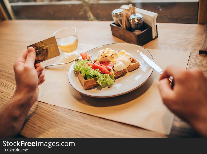 A picture of guy`s hands holding gold card in left one. He is holding a knife with right one. There is a plate with tasty food on table. Close up